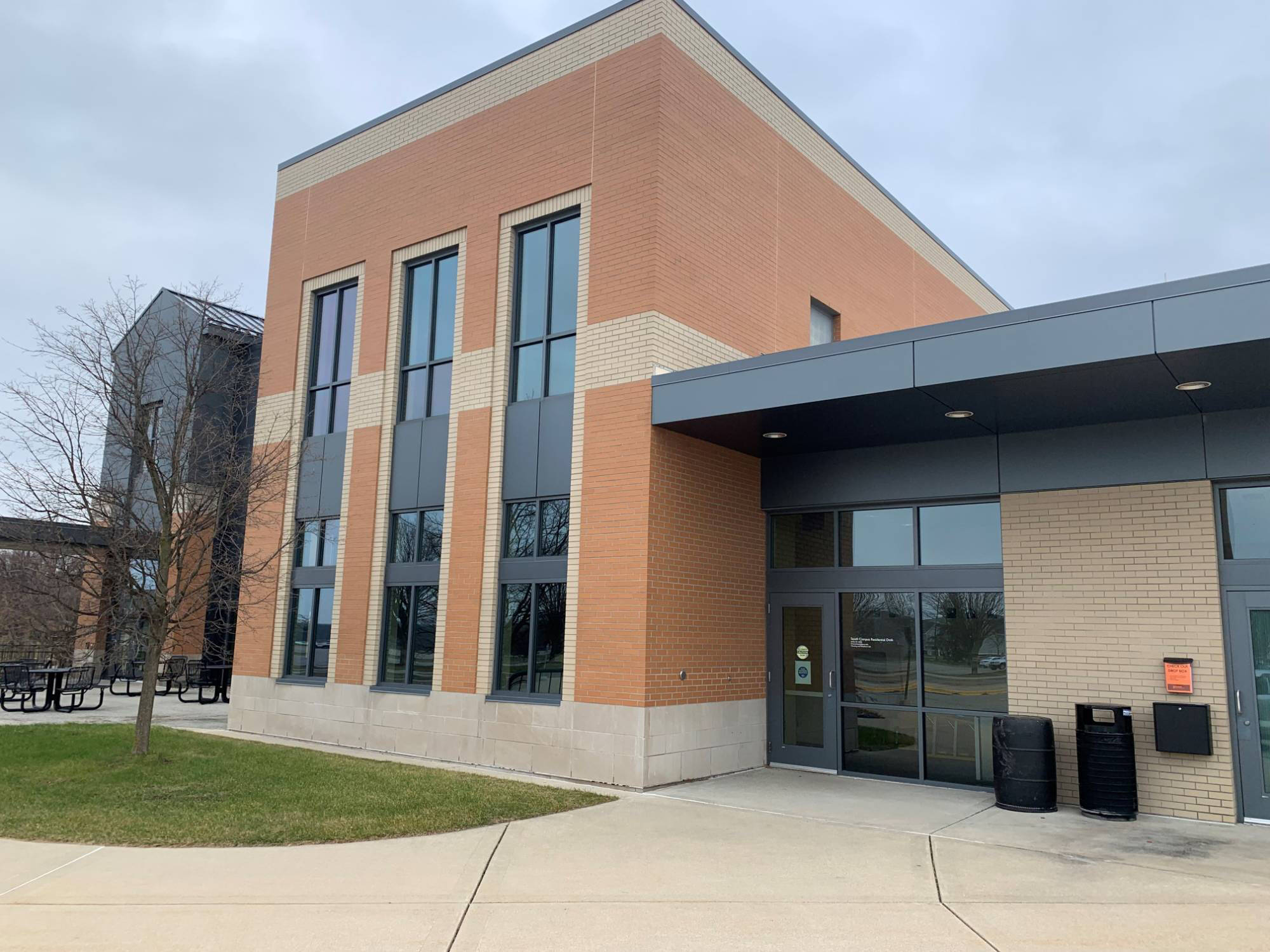 An orange, beige, and black building, where the South Campus Residential Desk is located, is surrounded by trees and a sidewalk leading up to the door.